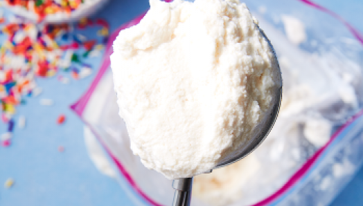 Gros plan d'une cuillère à crème glacée en argent avec une boule de glace à la vanille au-dessus d'un sac de glace à fermeture à glissière avec des paillettes colorées dans un bol sur la gauche.