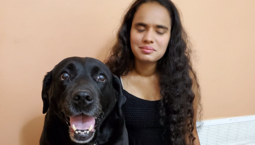 Runa Patel, kneeling on the floor next to her guide dog, smiling for the camera.