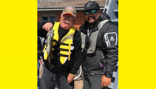 Bruce Roulston posant à côté de Eugene Chong avec un poisson pêché du quai sur la rive du Lake Joseph. Ils portent tous deux des gilets de sauvetage et une casquette. 