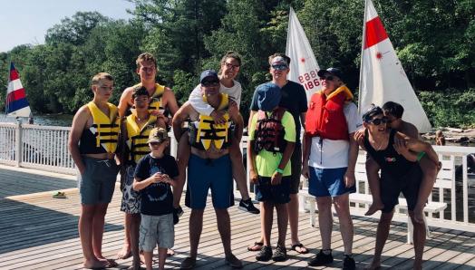 A group of people standing on the dock in shorts and life jackets. They are smiling and, in some cases, piggy-backing others.