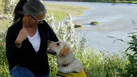 Debra Williamson kneeling next to Patsy, a yellow Labrador-Retriever wearing a Future Guide Dog vest, exchanging a glance with the other in front of the Bow River in Calgary