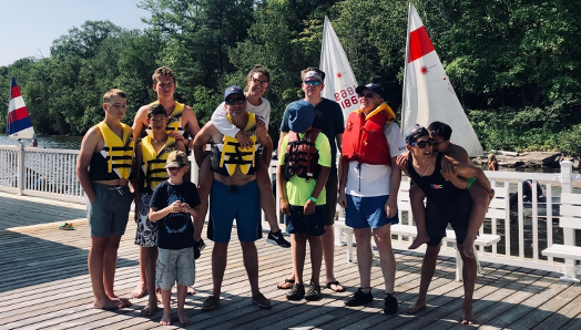 A small group of people standing on the dock in shorts and life jackets. They are smiling and, in some cases, piggy-backing others.