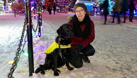 Lorraine Rempel kneeling on the snowy ground next to Jennie, a black Labrador-Retriever cross wearing a yellow Future Guide Dog vest. Trees decorated in lights are behind them.