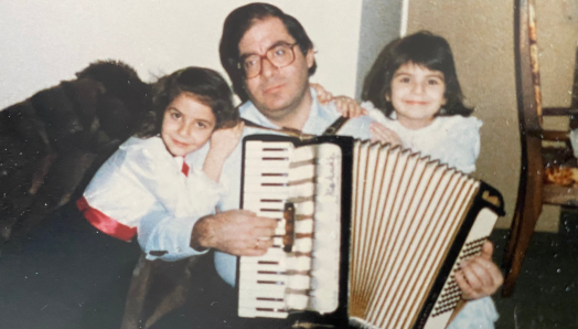 A photograph from the late ‘80s. Dimitrios plays the accordion. A young Angela sits on the left. Her sister sits on the right. 