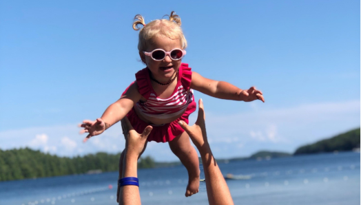 A little girl wearing a bathing suit and sunglasses is being thrown in the air by her mom on the shore at CNIB Lake Joe. 