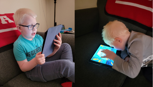 Lucas Burk (left) sits on a couch and holds his iPad with both hands. He is wearing glasses and a blue t-shirt. Ryan Burk (right) lays on a couch on his stomach and plays a game with his iPad. The iPad is leaning against the couch.
