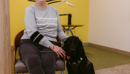 At a CNIB office, Penny sits in a chair. Her guide dog, Honour, a black Labrador retriever-golden retriever cross, sits at her feet. Penny pets Honour's head and smiles with joy.