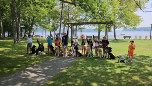 A group photo taken by the waterfront at CNIB Lake Joe. The group consists of participants with buddy dogs. They’re raising their hands in the air. 