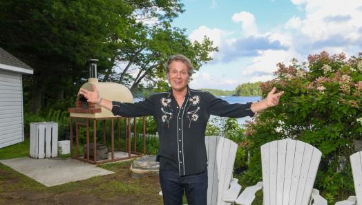 Jim Cuddy is standing in black shirt and jeans at the campfire circle in front of white Muskoka chairs and the outdoor pizza oven. He has his arms outstretched and is smiling.