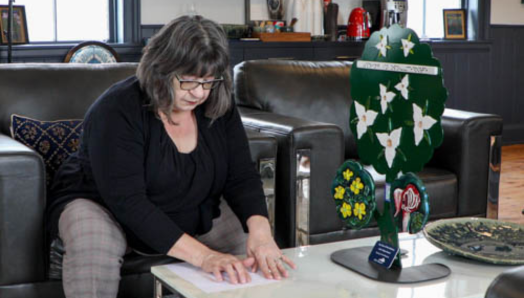 Robyn is sitting on a leather chair reading the Braille description of a tactile sculpture she was commissioned to make by Oro-Medonte Township. The sculpture is on the table and features tactile flowers: Trilliums, Marsh Marigolds, and a Lady’s Slipper.