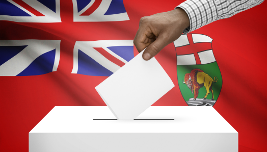 A hand dropping a ballot into a box; the Manitoba provincial flag appears in the background.