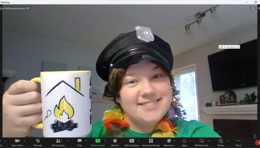 Sam is on a Zoom screen smiling. She is wearing a black police officer cap, a multi-colour Hawaiian lei, and is holding a CNIBLakeJoe@Home branded coffee mug.