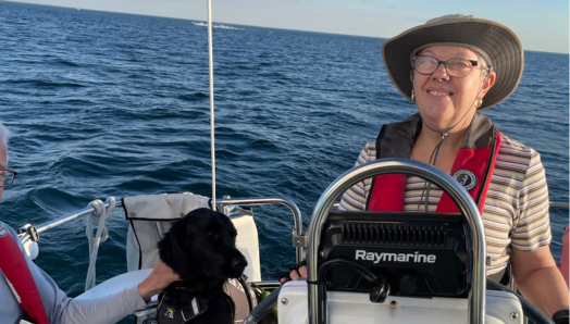 Sailing on Lake Ontario, Penny takes the helm of the boat. She is wearing glasses, a hat, and a red life jacket. Behind Penny is her black guide dog, Honour. To the left of Penny is a person who is partially visible, wearing a lifejacket. It’s a sunny day with a clear blue sky and a calm lake.  