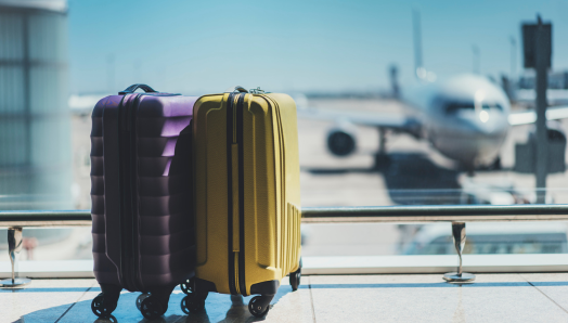 Two suitcases sit side-by-side in front of a window in an airport departure lounge. Outside the window, an airplane sits on the tarmac at the boarding gate. The airplane is out of focus and blurry.    